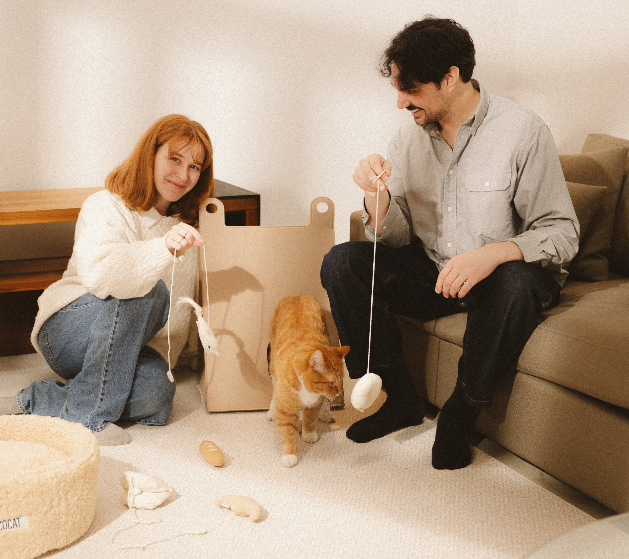 Two people playing with a cat on a beige couch in a living room.